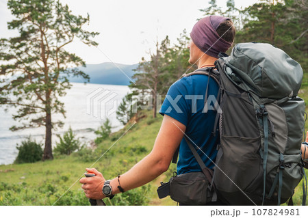 Hiking tourism adventure. Backpacker hiker man looking at beautiful view. Hiker tourist with backpack walking in green forest near lake. Back view man enjoy hike trekking tourism active vacation 107824981