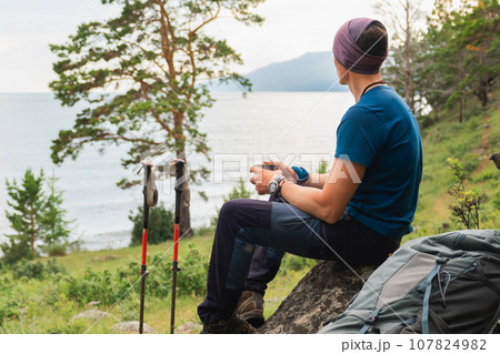 Hiking climbing cliff adventure. Backpacker man resting after hiking looking at beautiful view. Hiker with backpack sits on big rock above green forest and lake. Back view hiker man enjoy hike tourism 107824982