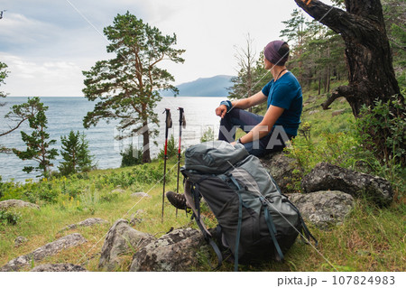 Hiking climbing cliff adventure. Backpacker man resting after hiking looking at beautiful view. Hiker with backpack sits on big rock above green forest and lake. Back view hiker man enjoy hike tourism 107824983