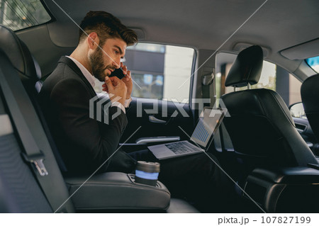 Busy entrepreneur working on laptop and talking on phone while going to airport by car 107827199