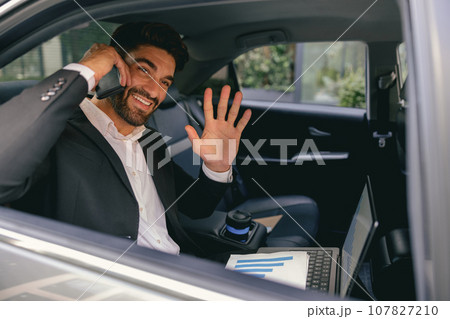 Smiling businessman in suit with laptop making phone call sit in taxi and waving Hi in open window Smiling businessman in suit with laptop making phone call sit in taxi and waving Hi in open window 107827210