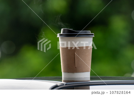 coffee takeaway in a paper cup on top of the car roof green tree background at sunrise in the morning,  selective focus, soft focus. 107828164