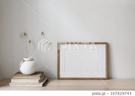 Horizontal blank wooden picture frame mockup on table. Trendy round ceramic vase with dry bunny tails, Lagurus ovatus grass and old books. White wall background. Scandinavian interior, home office. Horizontal blank wooden picture frame mockup on table. Trendy round ceramic vase with dry bunny tails, Lagurus ovatus grass and old books. White wall background. Scandinavian interior, home office. 107829743