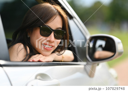 Attractive millennial woman in sunglasses leaning out of the car window enjoying the sunset. Active lifestyle, road trip and journey concept 107829748