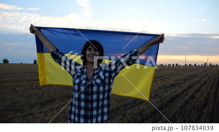 Happy woman looks into camera standing on wheat field with a lifted blue-yellow banner at sunset. Ukrainian smiling lady posing with a raised over head flag of Ukraine on barley meadow at sunrise. 107834039