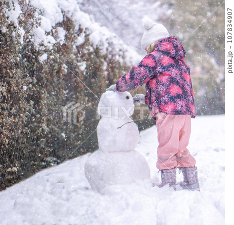 Little girl in warm winter clothes building snowman outdoors in park during snowfall. 107834907