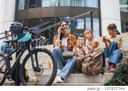 Children and mother have healthy snack in city after school. The family sitting on concrete steps in city, resting and eating home made snack, while mom is on the phone. 107837764