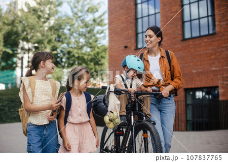 Mom picking up daughters from school with little son in bike seat . Schoolgirls telling mother about their day at school, walking in front of school building. 107837765