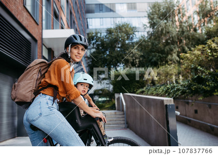 Mother carring her son on a child bike carrier, seat, both wearing helmets. Mom commuting with a young child through the city on a bicycle. Mother carring her son on a child bike carrier, seat, both wearing helmets. Mom commuting with a young child through the city on a bicycle. 107837766