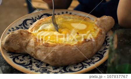 Close-up of hand of woman mixing suluguni cheese and egg yolk in Adjarian khachapuri, slow motion. Georgian traditional food concept Close-up of hand of woman mixing suluguni cheese and egg yolk in Adjarian khachapuri, slow motion. Georgian traditional food concept 107837767