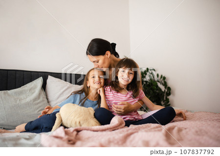 Mother with two daughters on a bed in the bedroom. Fatherless family sit and hug each other. Concept of Mother's Day and maternal love. 107837792
