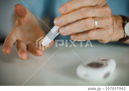 Diabetic senior man on wheelchair checking blood sugar level with fingerstick testing glucose meter. Elderly man with type 1 diabetes using blood glucose monitor at home. 107837942