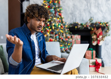Upset man looking at laptop screen sitting at home in living room on Christmas, hispanic man frustrated writing online notification email to mail, near Christmas tree on New Year holidays. 107838482
