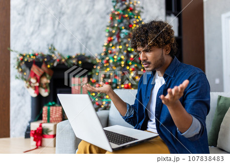 Upset man looking at laptop screen sitting at home in living room on Christmas, hispanic man frustrated writing online notification email to mail, near Christmas tree on New Year holidays. 107838483