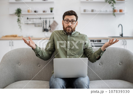 Confused male shrugging shoulders with laptop on couch 107839330