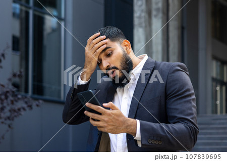 Upset Muslim male businessman standing near office and holding head, shocked reading news, message received. 107839695