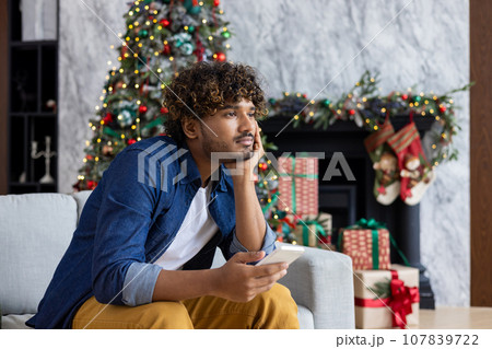 Bored man sitting on sofa for New Year holidays and Christmas, yawning hispanic man holding smartphone in living room near Christmas tree. 107839722