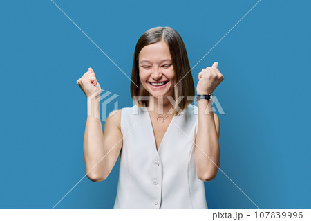 Young cheerful woman clenching her fists on blue background Young cheerful woman clenching her fists on blue background 107839996