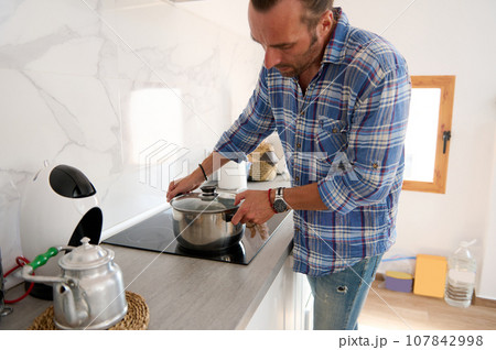 Young adult Caucasian handsome young man putting a saucepan on an electric stove while cooking dinner in home kitchen Young adult Caucasian handsome young man putting a saucepan on an electric stove while cooking dinner in home kitchen 107842998