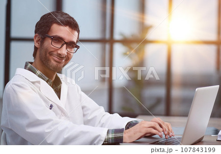 A handsome young doctor sits at a table and smiles friendly while looking at the camera. A handsome young doctor sits at a table and smiles friendly while looking at the camera. 107843199