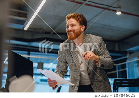Smiling businessman holding documents and looks away while standing in office 107846625