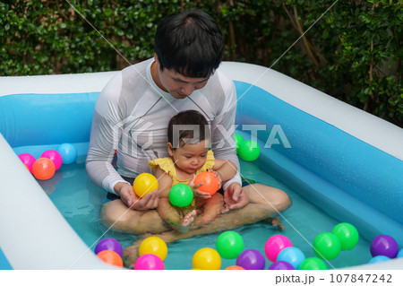 father and infant baby girl playing water with colorful plastic balls in inflatable pool 107847242