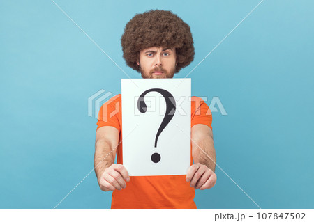 Portrait of man with Afro hairstyle wearing orange T-shirt holding question mark, finding smart solution, asking for advice, looking at camera. Indoor studio shot isolated on blue background. Portrait of man with Afro hairstyle wearing orange T-shirt holding question mark, finding smart solution, asking for advice, looking at camera. Indoor studio shot isolated on blue background. 107847502