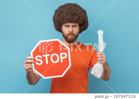 Portrait of serious responsible man with Afro hairstyle wearing orange T-shirt holding red stop sign and plastic package, looking at camera. Indoor studio shot isolated on blue background. 107847519