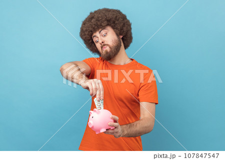Portrait of man with Afro hairstyle wearing orange T-shirt investing, putting dollar banknote into piggy bank, proud with money savings. Indoor studio shot isolated on blue background. Portrait of man with Afro hairstyle wearing orange T-shirt investing, putting dollar banknote into piggy bank, proud with money savings. Indoor studio shot isolated on blue background. 107847547
