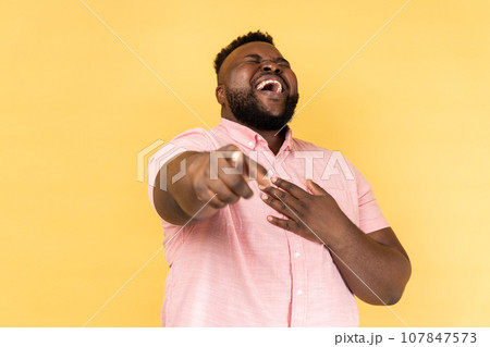 Portrait of funny positive bearded young adult man wearing pink shirt laughing out loud holding belly and pointing finger on you, mockery. Indoor studio shot isolated on yellow background. Portrait of funny positive bearded young adult man wearing pink shirt laughing out loud holding belly and pointing finger on you, mockery. Indoor studio shot isolated on yellow background. 107847573