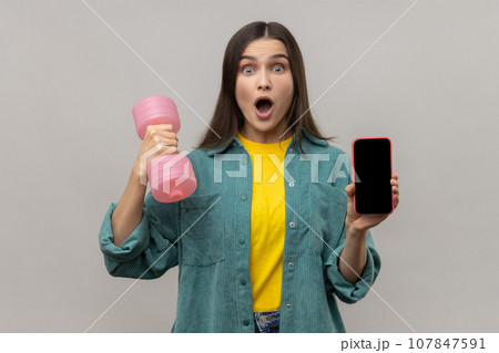 Portrait of amazed surprised holding pink dumbbell and blank screen smartphone in hands, satisfied with sports app, wearing casual style jacket. Indoor studio shot isolated on gray background. 107847591