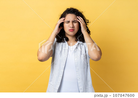 Portrait of woman with dark wavy hair frowning and clasping sore head, suffering intense headache, having migraine, fever symptoms. Indoor studio shot isolated on yellow background. 107847600