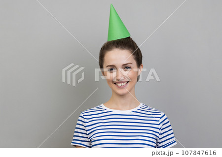 Portrait of smiling positive delighted woman wearing striped T-shirt and party cone, looking at camera with toothy smile. Indoor studio shot isolated on gray background. 107847616