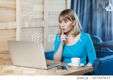 Portrait of serious adorable young woman with blonde hair in blue shirt sitting at table and working on laptop, has video call, showing shh gesture. Indoor shot, cafe background. Portrait of serious adorable young woman with blonde hair in blue shirt sitting at table and working on laptop, has video call, showing shh gesture. Indoor shot, cafe background. 107847624