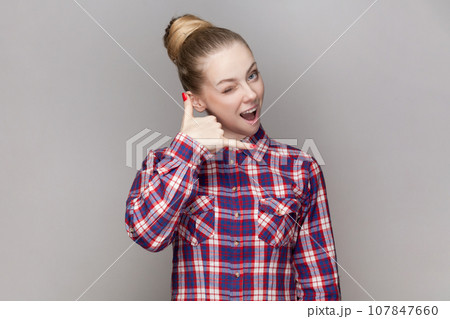 Portrait of beautiful positive woman with bun hairstyle flirting with attractive guy, showing call me gesture, wearing checkered shirt. Indoor studio shot isolated on gray background. Portrait of beautiful positive woman with bun hairstyle flirting with attractive guy, showing call me gesture, wearing checkered shirt. Indoor studio shot isolated on gray background. 107847660