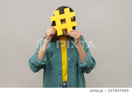Portrait of curious woman looking through yellow hashtag sign, looking for proper posts in social media, spying, wearing casual style jacket. Indoor studio shot isolated on gray background. Portrait of curious woman looking through yellow hashtag sign, looking for proper posts in social media, spying, wearing casual style jacket. Indoor studio shot isolated on gray background. 107847668