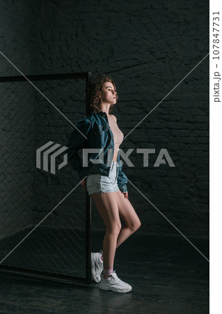 Full length profile portrait of beautiful young adult woman with wavy hair standing near fence on dark brick wall background, looking away with serious expression. Indoor studio shot. 107847731