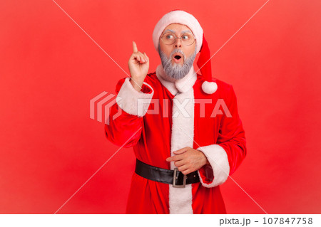 Elderly man with gray beard wearing santa claus costume raising finger and having genius idea of Christmas celebration, looking amazed, creative thought. Indoor studio shot isolated on red background. Elderly man with gray beard wearing santa claus costume raising finger and having genius idea of Christmas celebration, looking amazed, creative thought. Indoor studio shot isolated on red background. 107847758