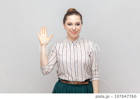 Portrait of pleased pretty positive woman wearing striped shirt and green skirt standing waving hand, saying hello or bye, having meeting. Indoor studio shot isolated on gray background. Portrait of pleased pretty positive woman wearing striped shirt and green skirt standing waving hand, saying hello or bye, having meeting. Indoor studio shot isolated on gray background. 107847760