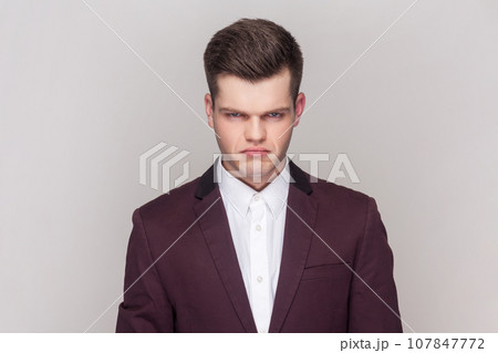 Portrait of gloomy sad handsome man has unhappy look, expresses regret and sadness, frowns face, being frustrated, wearing violet suit and white shirt. Indoor studio shot isolated on grey background. 107847772