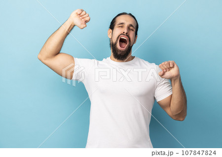 Portrait of sleepy handsome man with beard wearing white T-shirt standing and yawning with closed eyes and raised arms, waking up early. Indoor studio shot isolated on blue background. Portrait of sleepy handsome man with beard wearing white T-shirt standing and yawning with closed eyes and raised arms, waking up early. Indoor studio shot isolated on blue background. 107847824