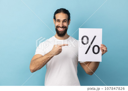 Portrait of attractive optimistic man with beard wearing white T-shirt holding and pointing paper with percent sign inscription, looking at camera. Indoor studio shot isolated on blue background. Portrait of attractive optimistic man with beard wearing white T-shirt holding and pointing paper with percent sign inscription, looking at camera. Indoor studio shot isolated on blue background. 107847826