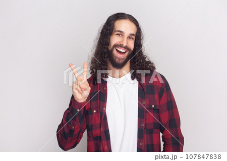 Portrait of optimistic glad bearded man with long curly hair in checkered red shirt with positive facial expression, shows V sign or victory gesture. Indoor studio shot isolated on gray background. 107847838