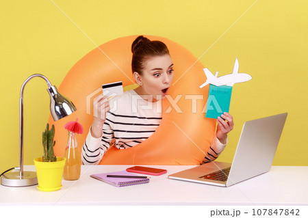 Amazed shocked woman with rubber ring holding and showing credit card and passport, looking at laptop display with open mouth. Indoor studio studio shot isolated on yellow background. Amazed shocked woman with rubber ring holding and showing credit card and passport, looking at laptop display with open mouth. Indoor studio studio shot isolated on yellow background. 107847842