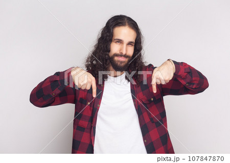 Portrait of bossy anger bearded man with long curly hair in checkered red shirt pointing down and looking arrogant, demanding to do immediately. Indoor studio shot isolated on gray background. Portrait of bossy anger bearded man with long curly hair in checkered red shirt pointing down and looking arrogant, demanding to do immediately. Indoor studio shot isolated on gray background. 107847870