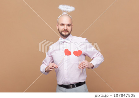 Portrait of cute friendly bald bearded man with nimb over head, holding little red hearts in front of his chest, wearing light pink shirt and bow tie. Indoor studio shot isolated on brown background. Portrait of cute friendly bald bearded man with nimb over head, holding little red hearts in front of his chest, wearing light pink shirt and bow tie. Indoor studio shot isolated on brown background. 107847908