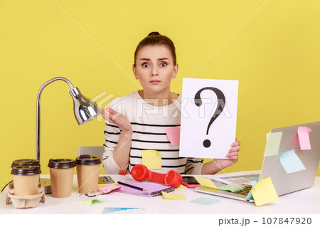 Portrait of shocked astonished woman looking at camera, holding paper with question mark, thinks about tasks, covered with sticky notes. Indoor studio studio shot isolated on yellow background. 107847920