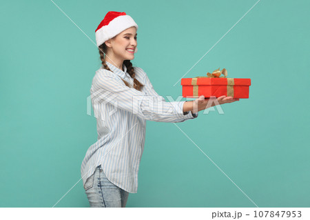 Side view portrait of smiling delighted teenager girl wearing striped shirt and Santa Claus hat, congratulating with new year, giving present. Indoor studio shot isolated on green background. Side view portrait of smiling delighted teenager girl wearing striped shirt and Santa Claus hat, congratulating with new year, giving present. Indoor studio shot isolated on green background. 107847953