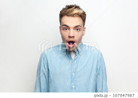 Portrait of amazed surprised man wearing denim shirt looking at camera with big eyes and open mouth, sees something shocked and astonished. Indoor studio shot isolated on gray background. Portrait of amazed surprised man wearing denim shirt looking at camera with big eyes and open mouth, sees something shocked and astonished. Indoor studio shot isolated on gray background. 107847987