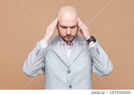 Portrait of sick ill unhealthy bald bearded man standing with hands on head, keeps eyes closed, suffering terrible headache, wearing gray jacket. Indoor studio shot isolated on brown background. 107848000